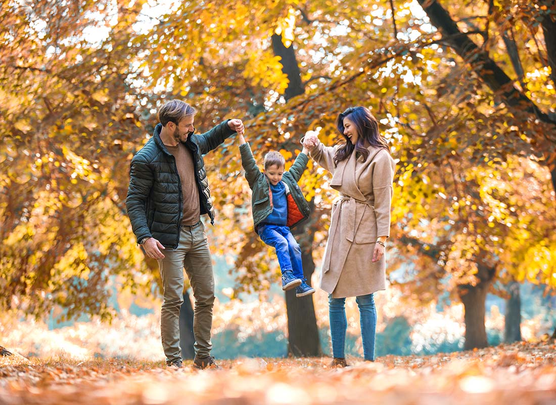 About Our Agency - Portrait of Two Joyful Parents Holding Up Their Son in the Air as They Walk Through a Park During the Fall