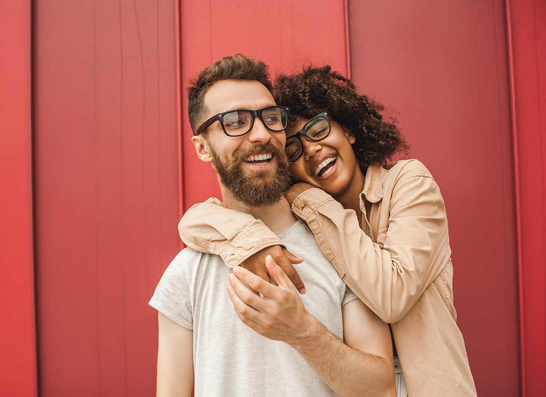 Read Our Reviews - Portrait of a Young Cheerful Couple Wearing Glasses Hugging Each Other as They Stand Outside a Red Building