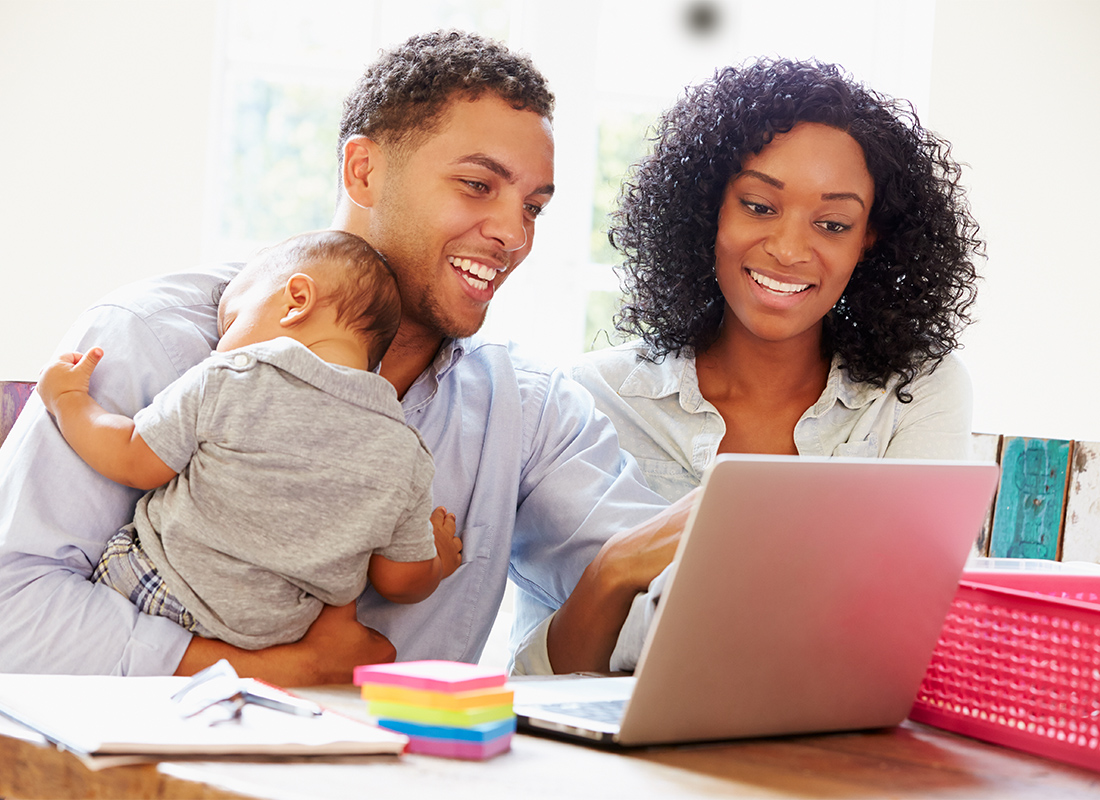 Frequently Asked Questions - Smiling Mother and Father Holding a Child While Looking at an Open Laptop in their Home with Light Pouring through the Window Behind Them
