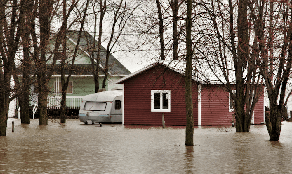 Flooded residential area with water surrounding a red home and camper, highlighting the risks of spring flooding and the need for flood insurance coverage.
