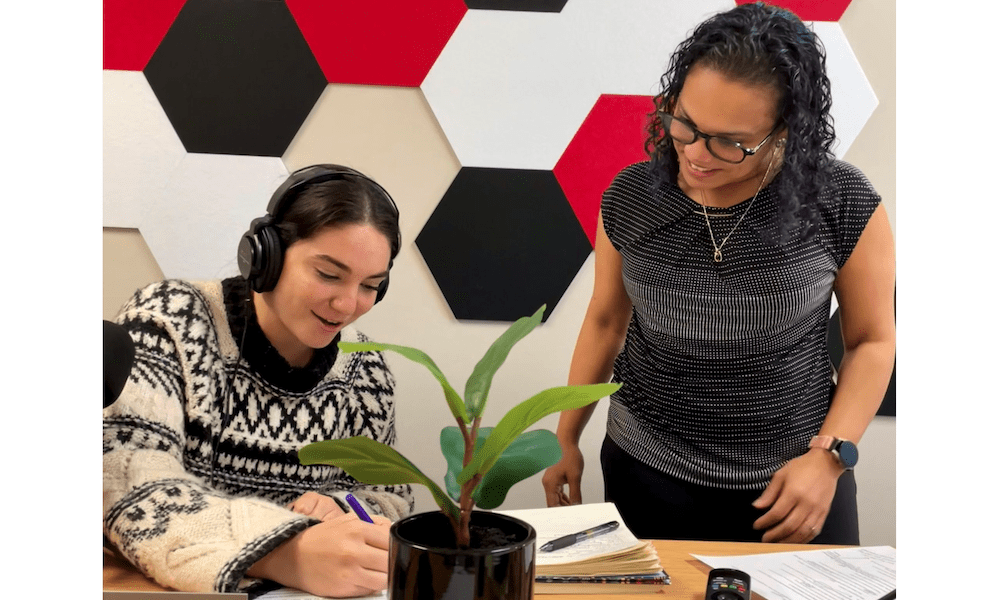 Casandra Magliane mentoring a YMI Insurance employee in the podcast room, as the employee listens with headphones and takes notes.