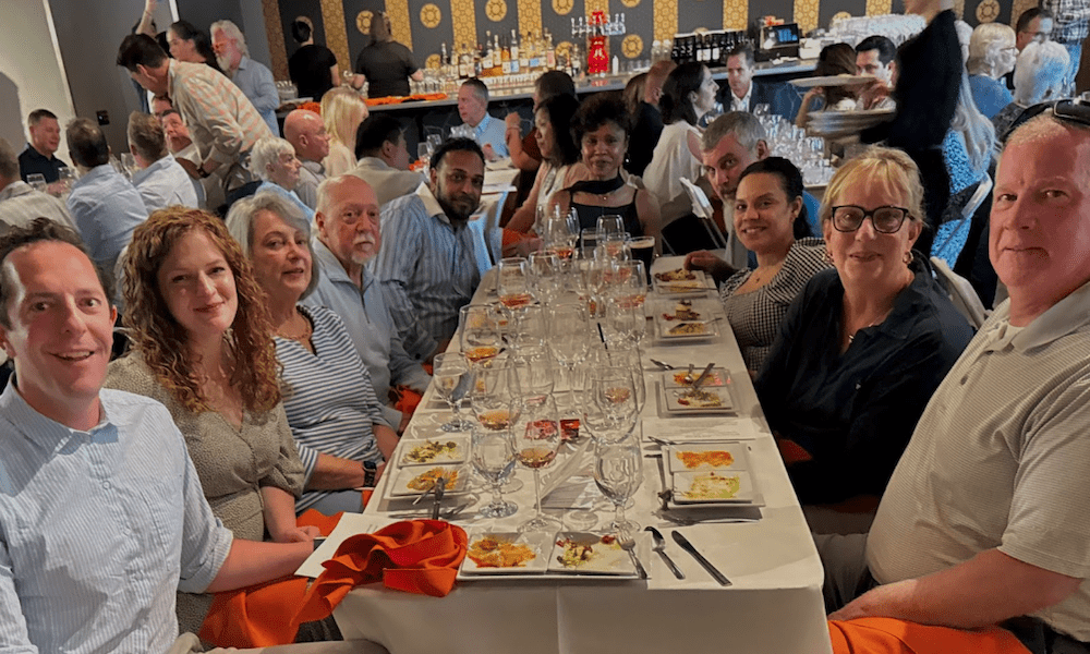 The YMI Insurance management team seated at a long, elegantly set dinner table during the Wheels of Thyme fundraiser at Mister Lee’s Noodles. The group is smiling, surrounded by tasting plates, empty wine glasses, and a lively crowd in the background.