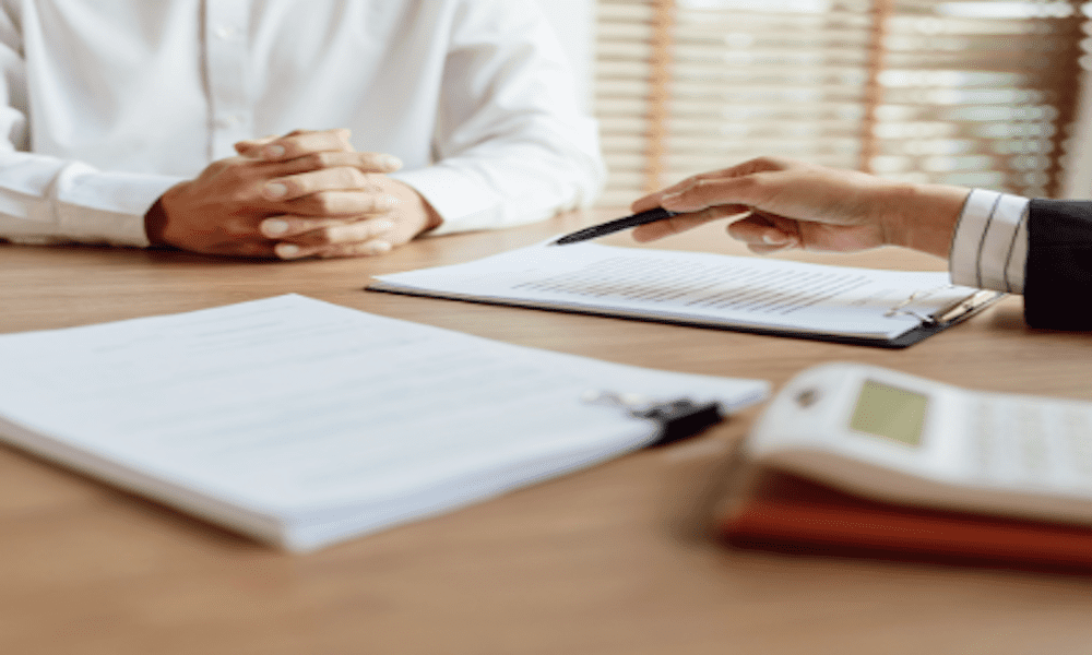 Business insurance agent discussing policy details with a client at a desk, with documents, a calculator, and contract paperwork in a professional office setting.