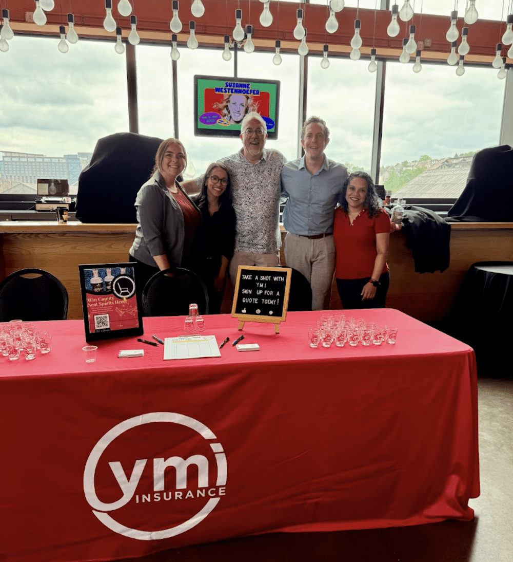 Jay Nee and the YMI team smiling in front of the YMI-sponsored table at the 2025 Ginvitational at ArtsQuest. The table is decorated with branded shot glasses and signage reading “Take a shot with YMI”.