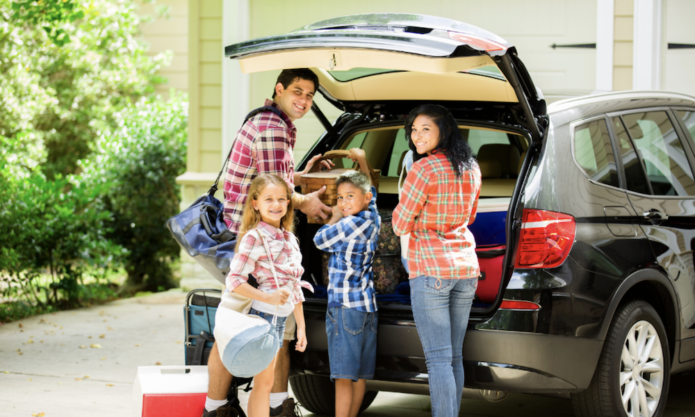A smiling family of four stands behind an SUV, packing for a trip. The parents and two children, all dressed casually in plaid shirts, are loading bags and a cooler into the open trunk in a sunny driveway, preparing for vacation.