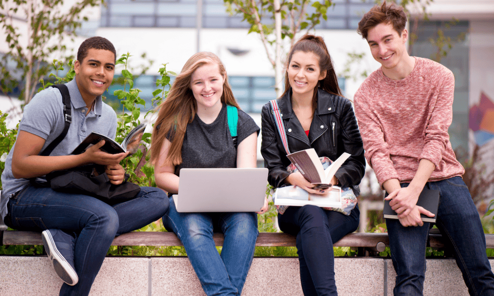 Four college students sit outside on a campus bench, smiling and studying together. One student uses a laptop, while the others hold books and notebooks. Trees and a modern academic building are visible in the background.