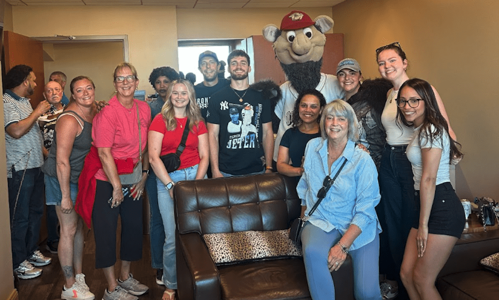 The YMI team poses with the IronPigs mascot inside a private suite at Coca-Cola Park in Allentown, PA during a summer baseball game. Team members from Stroudsburg, Bethlehem, and Allentown gathered for this company outing, hosted by Metro Decorators—an interior design company located in the Poconos. The box features leather couches and warm lighting, and the group is smiling and enjoying a fun team-building night out at the stadium.