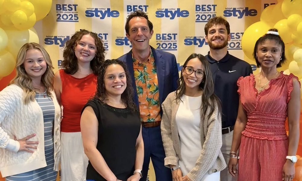 A group of eight YMI Insurance team members smiling and posing in front of a yellow and white “Best of the Lehigh Valley 2025” step-and-repeat banner. The backdrop is framed with yellow and orange balloon arrangements, and everyone is dressed in business casual attire.