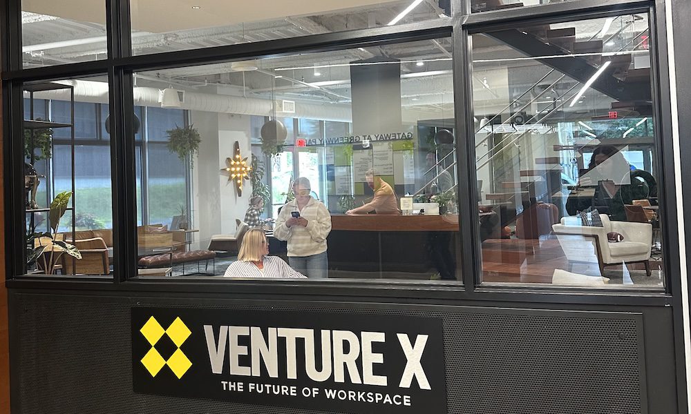 Front view of Venture X Bethlehem coworking space, showing large glass windows, modern industrial-style interior with plants and seating, and a bold black-and-yellow Venture X logo sign that reads “The Future of Workspace.”