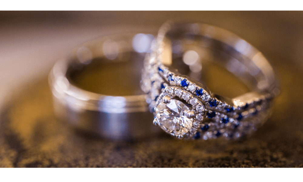 A Love Story Worth Protecting/ How to Insure Your Engagement Ring in the Lehigh Valley - Close-up of diamond engagement ring with sapphire accents next to a silver wedding band on a rustic surface