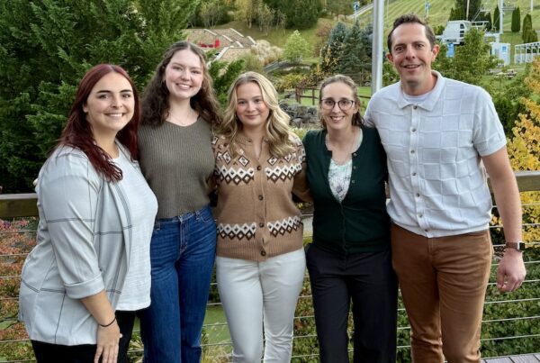 Five YMI team members standing together outdoors at Bear Creek Mountain Resort during the 2025 IA&B Young Agents Conference. The group is smiling on a deck with a scenic backdrop of trees and ski slopes in early fall.