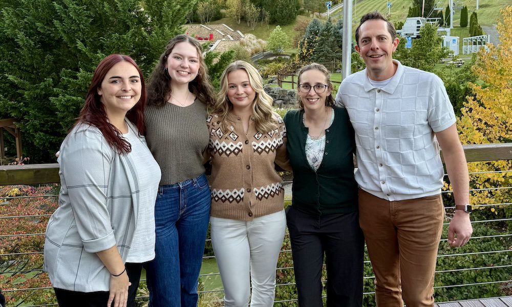 Five YMI team members standing together outdoors at Bear Creek Mountain Resort during the 2025 IA&B Young Agents Conference. The group is smiling on a deck with a scenic backdrop of trees and ski slopes in early fall.