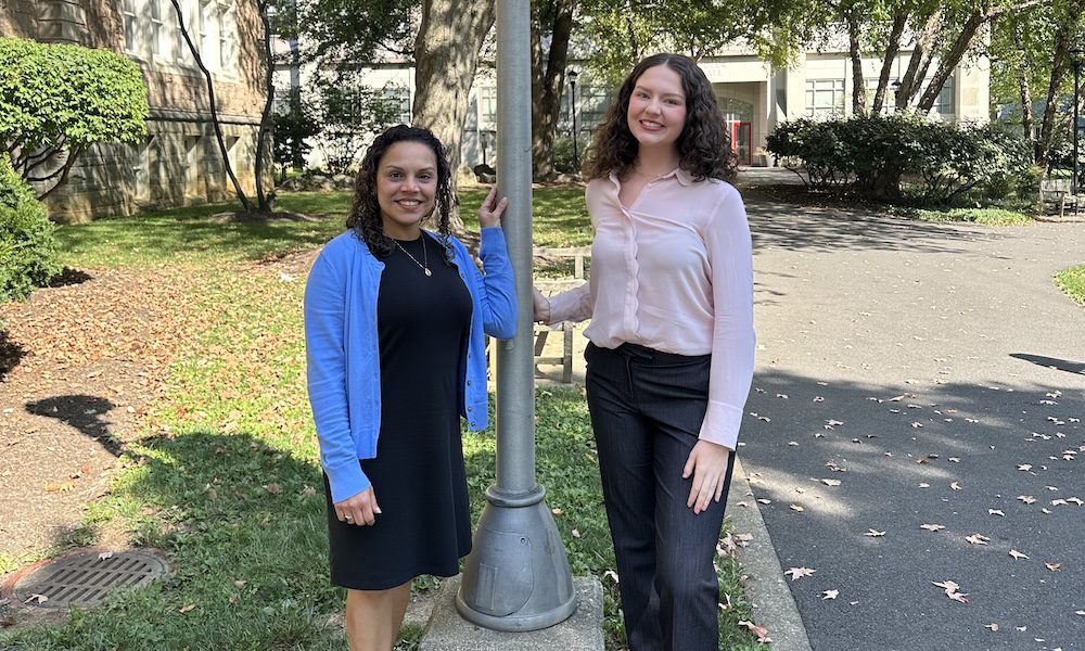 "Emily Ellis and Casandra Magliane from YMI Agency standing outside at Muhlenberg College in Allentown, PA, smiling beside a red campus banner on a sunny day."