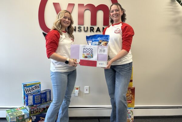Two YMI team members stand in front of the YMI Insurance logo holding a donation box for the Northeast Community Center Food Drive, surrounded by collected non-perishable food items like pasta, canned goods, and cereal.”