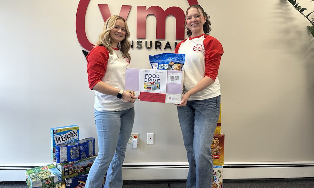 Two YMI team members stand in front of the YMI Insurance logo holding a donation box for the Northeast Community Center Food Drive, surrounded by collected non-perishable food items like pasta, canned goods, and cereal.”