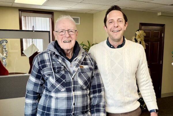 Alt text: Two men stand smiling inside an insurance office decorated for the holidays. An older man wearing glasses, a blue plaid jacket, and jeans stands on the left holding a camouflage hat. A younger man in a cream-colored cable-knit sweater and brown pants stands beside him on the right. Behind them are chairs, stockings hung near a divider, and warm, neutral office decor.