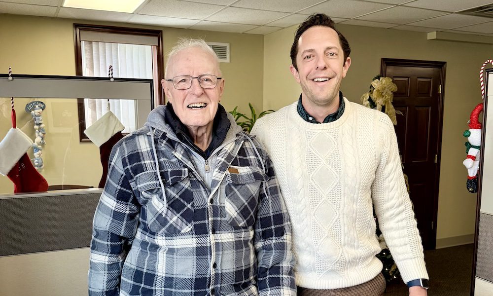 Alt text: Two men stand smiling inside an insurance office decorated for the holidays. An older man wearing glasses, a blue plaid jacket, and jeans stands on the left holding a camouflage hat. A younger man in a cream-colored cable-knit sweater and brown pants stands beside him on the right. Behind them are chairs, stockings hung near a divider, and warm, neutral office decor.