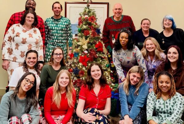 Group photo of the YMI team wearing festive holiday pajamas, gathered around a decorated Christmas tree in an office setting, smiling and celebrating together.