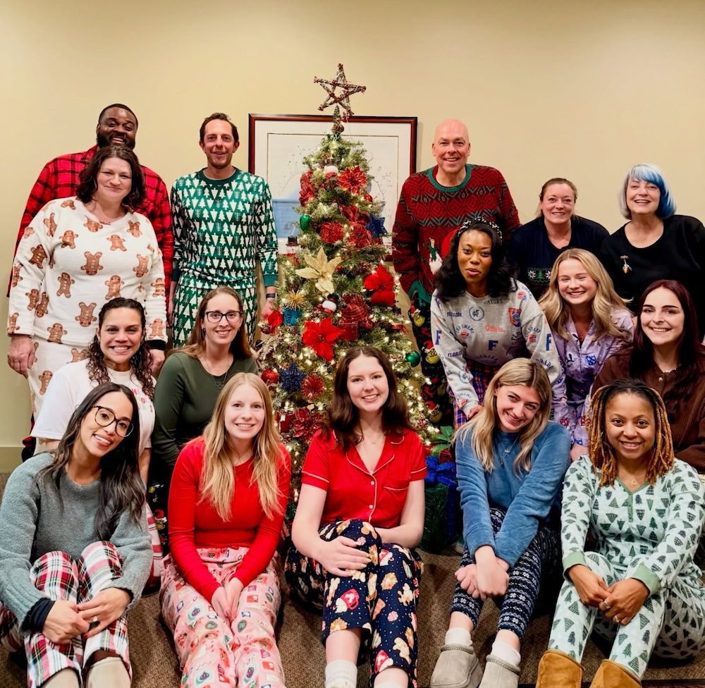 Group photo of the YMI team wearing festive holiday pajamas, gathered around a decorated Christmas tree in an office setting, smiling and celebrating together.