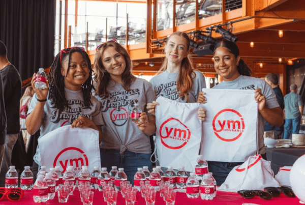 Four YMI team members pose behind the YMI giveaway table at the Ginvitational event at SteelStacks, showcasing branded drawstring bags, water bottles, sunglasses, shot glasses, and beach balls while welcoming attendees.
